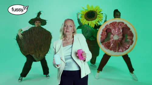 Playful studio scene with Deborah Meaden from Dragon's Den holding two Fussy deodorant cases, standing in front of three people dressed in oversized fruit and plant costumes 