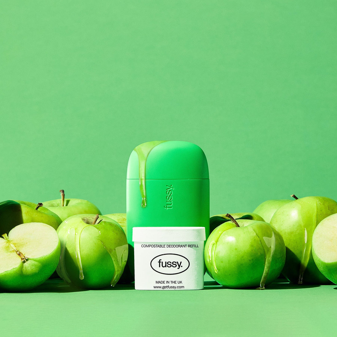 Fussy's Toffee Apple product display featuring a reusable deodorant case surrounded by fresh green apples, with syrup drips, and a refill pack placed in front against a matching green background