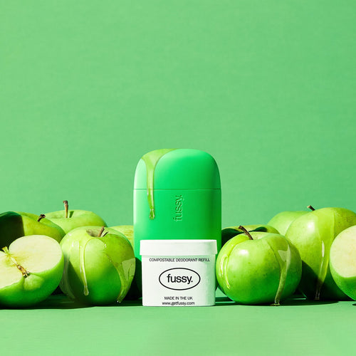 Fussy's Toffee Apple product display featuring a reusable deodorant case surrounded by fresh green apples, with syrup drips, and a refill pack placed in front against a matching green background