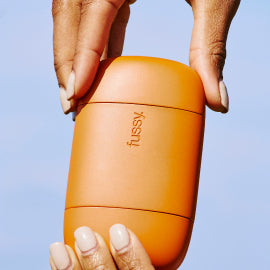 Orange Fussy deodorant case held up to a blue sky